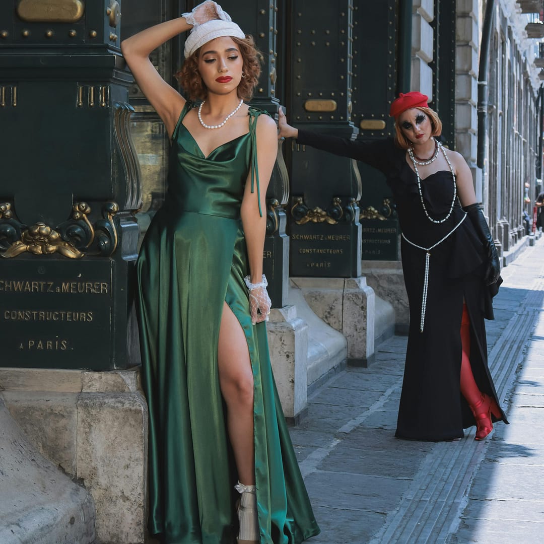 Two women pose stylishly on a city street. One wears a green dress, white gloves and white beret while the other complements her black and red outfit with a chic red beret. They stand in front of ornate doors