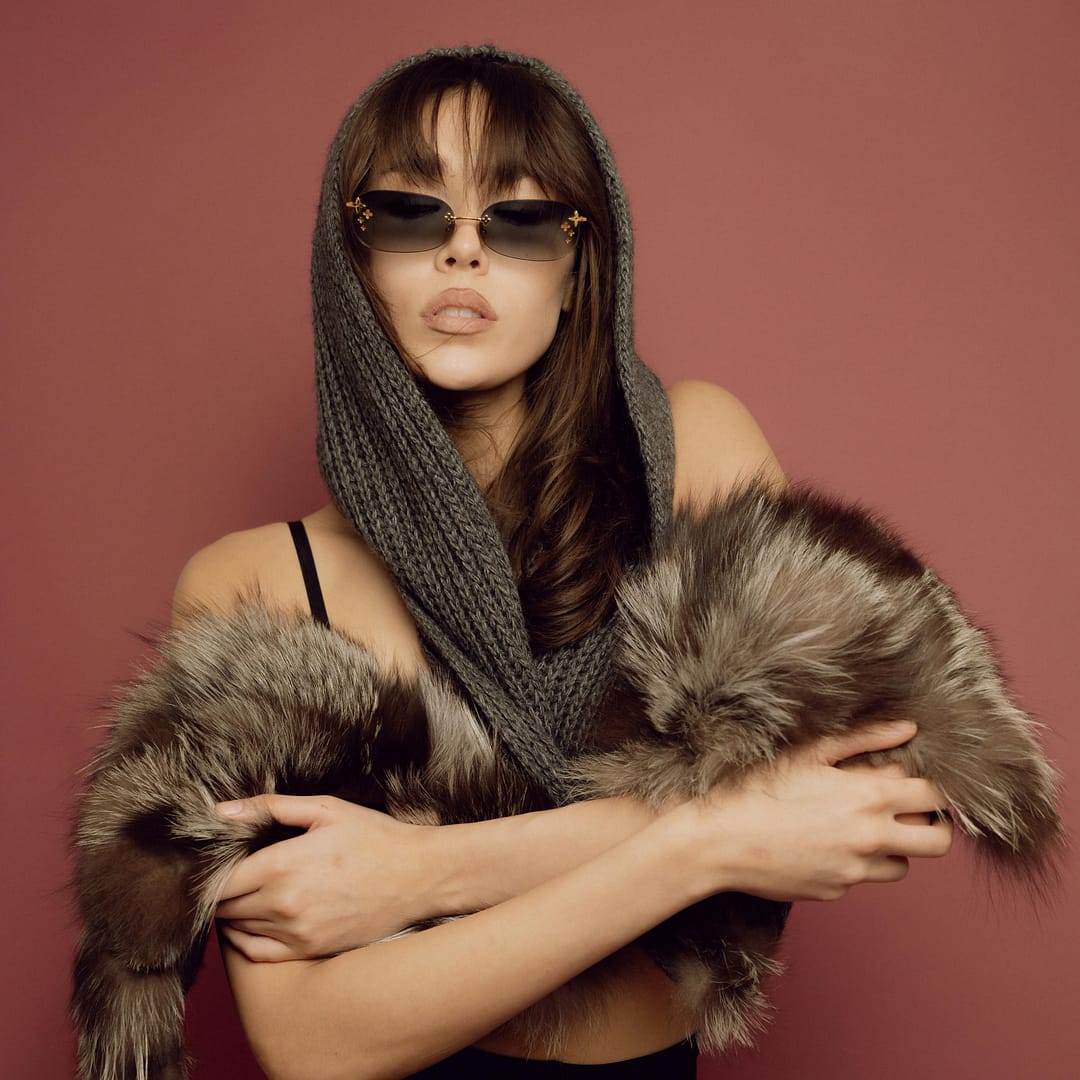 Woman wearing sunglasses and a knitted hood poses against a reddish background, draped in a faux fur garment