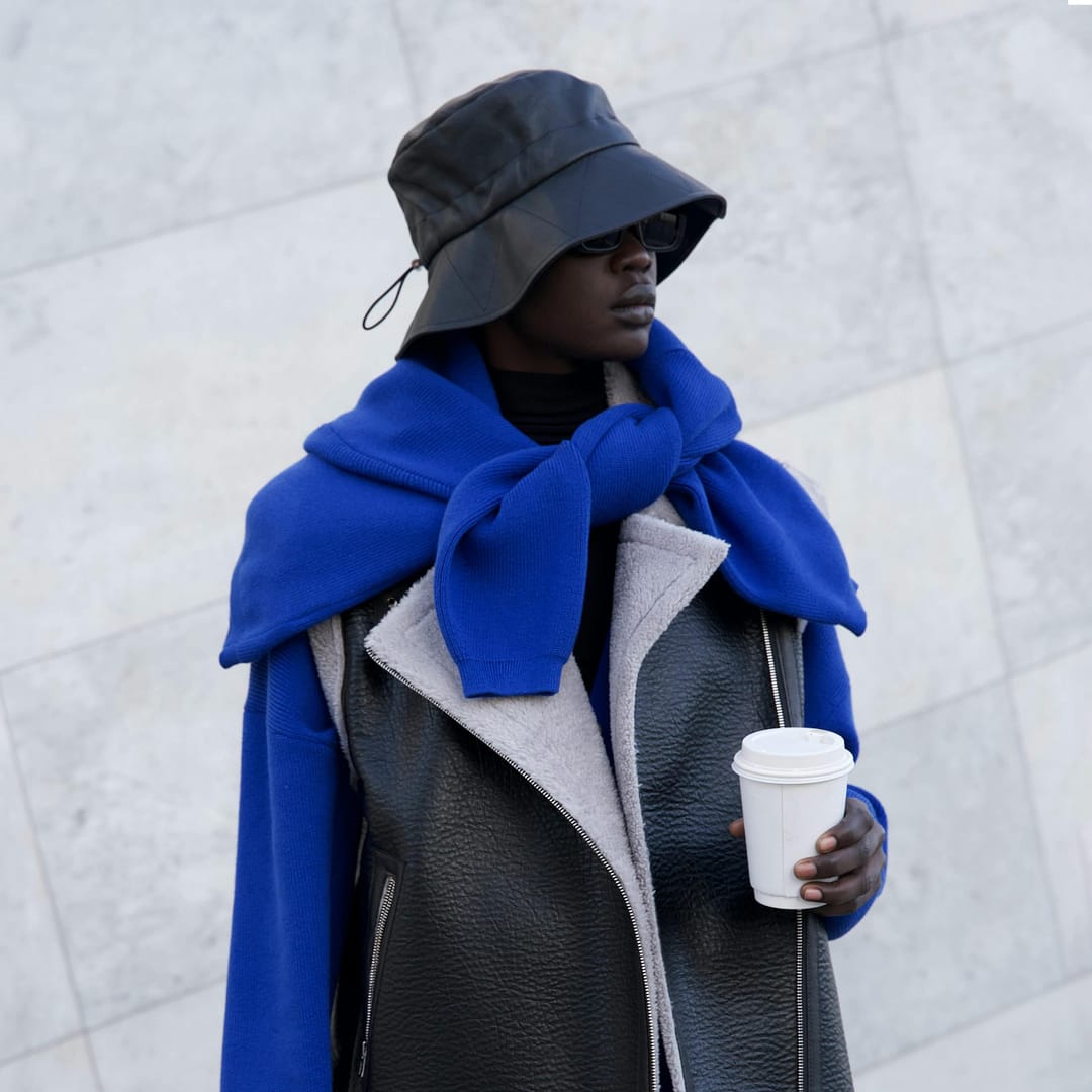 Black woman in a black leather bucket hat, blue sweater, and leather vest holds a coffee cup while standing against a light-colored wall