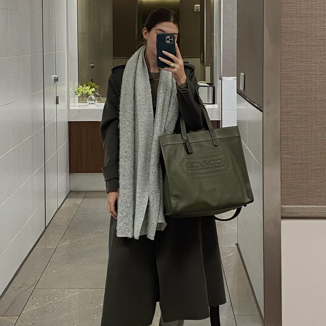 Person in a mirror selfie wearing a long dark coat, gray scarf, and sneakers, holding a large green Coach designer tote bag, in a modern hallway with a tiled wall and flowers in the background