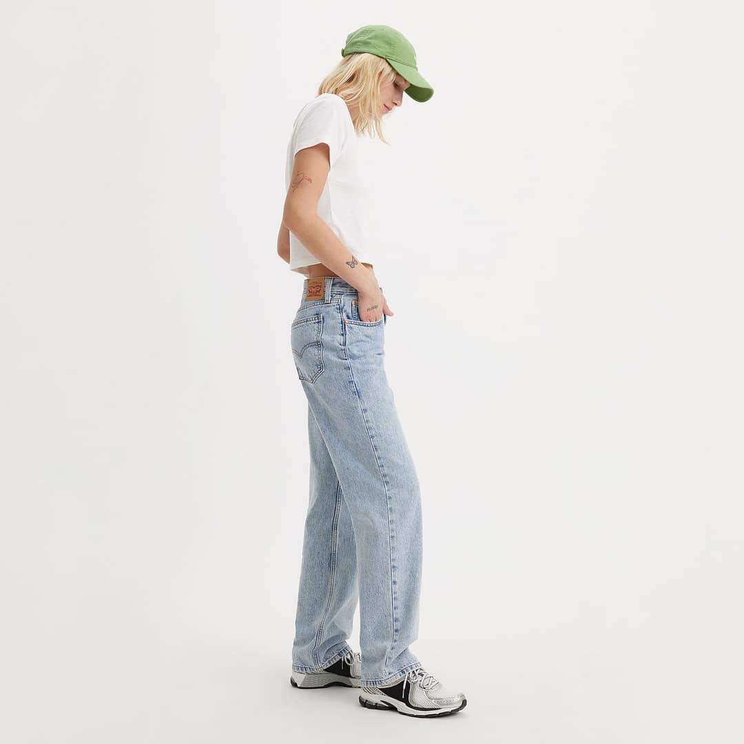 Woman wearing a white shirt, low-rise light blue jeans, a green cap, and sneakers stands sideways against a plain background