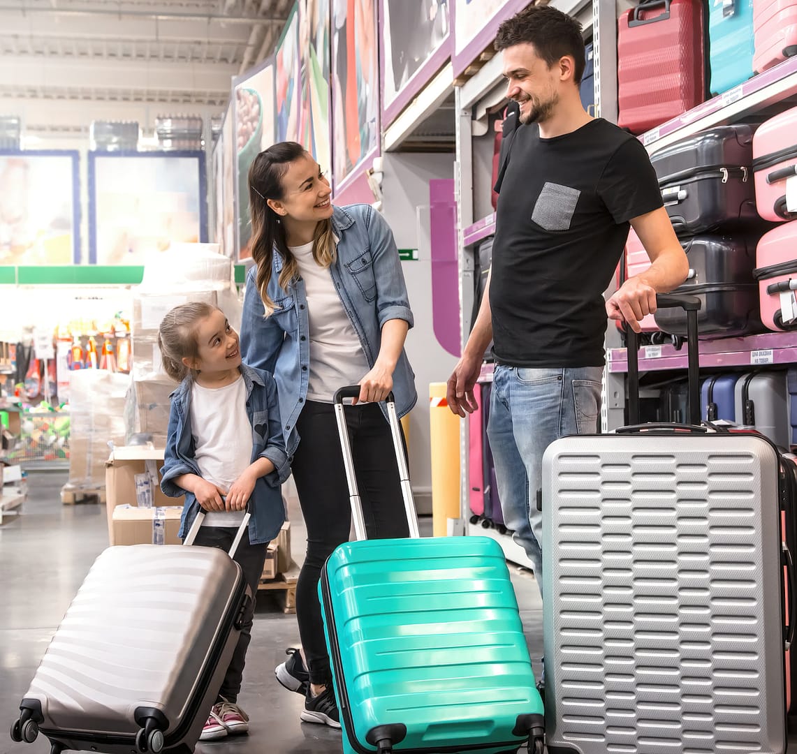 A family of three, consisting of a couple and their young daughter, shopping for luggage in a store aisle, smiling and chatting together