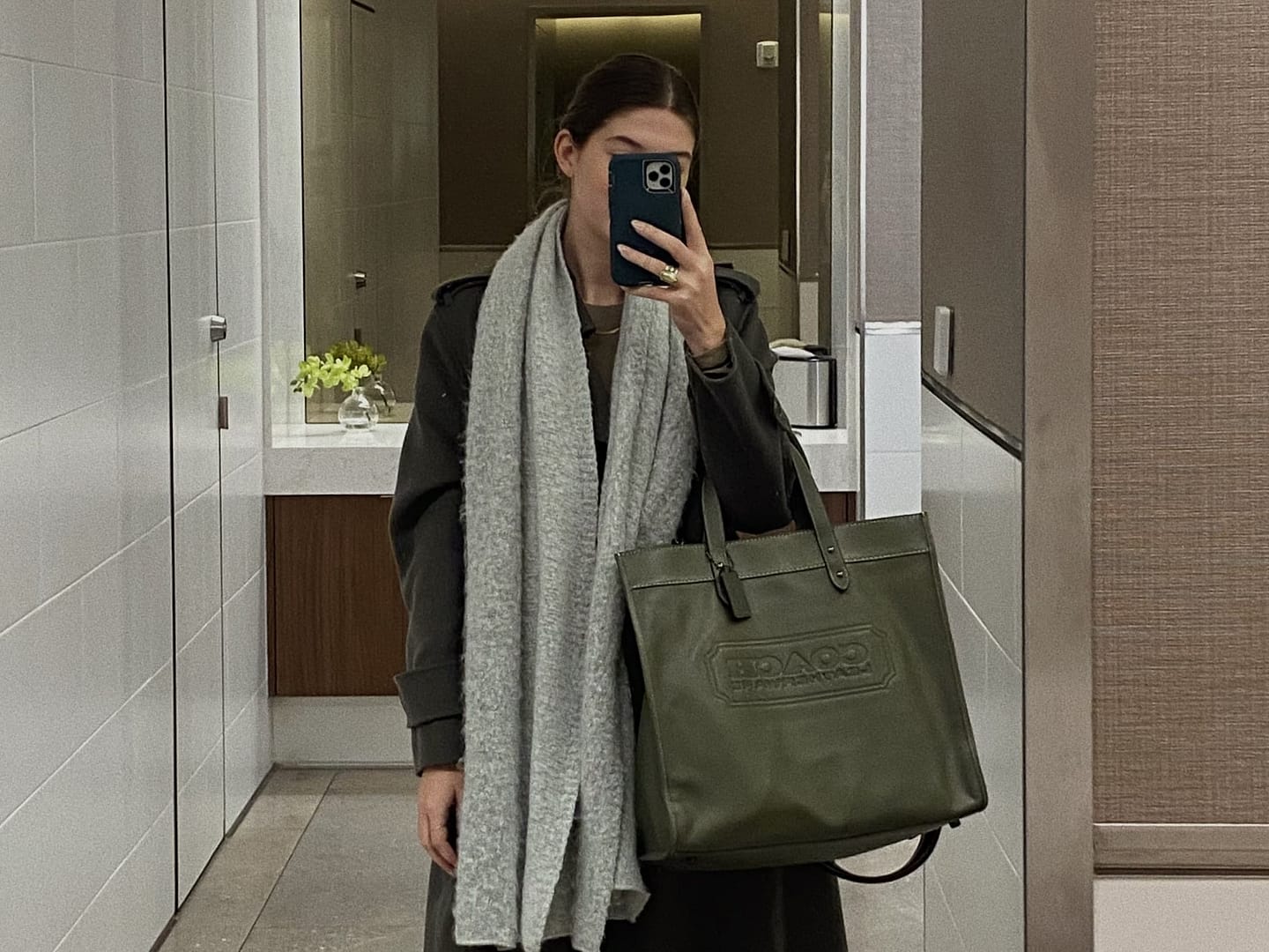 Person in a mirror selfie wearing a long dark coat, gray scarf, and sneakers, holding a large green Coach designer tote bag, in a modern hallway with a tiled wall and flowers in the background