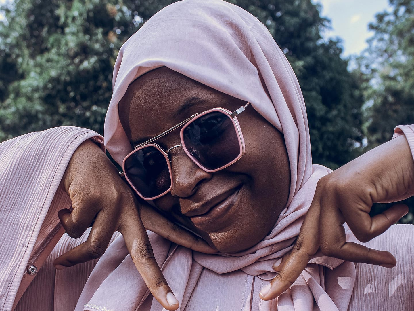 Black woman wearing a pink hijab and stylish sunglasses is smiling warmly while making a friendly hand gesture outdoors