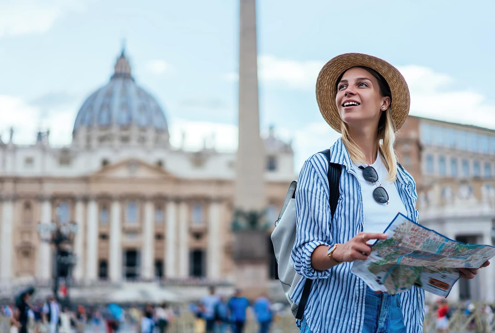Smiling female tourist in straw hat enjoying beautiful architecture in Vatican feeling carefree during recreation journey, happy Caucasian hipster girl holding map having sightseeing tour near Rome