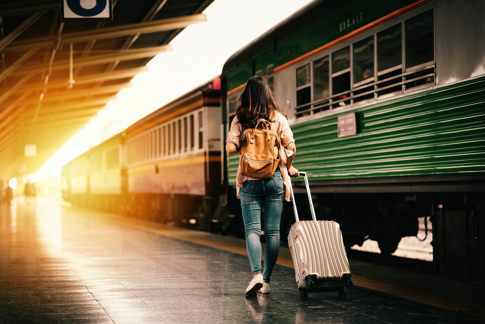 Woman with a backpack pulling a suitcase walks towards a train at a station during sunset