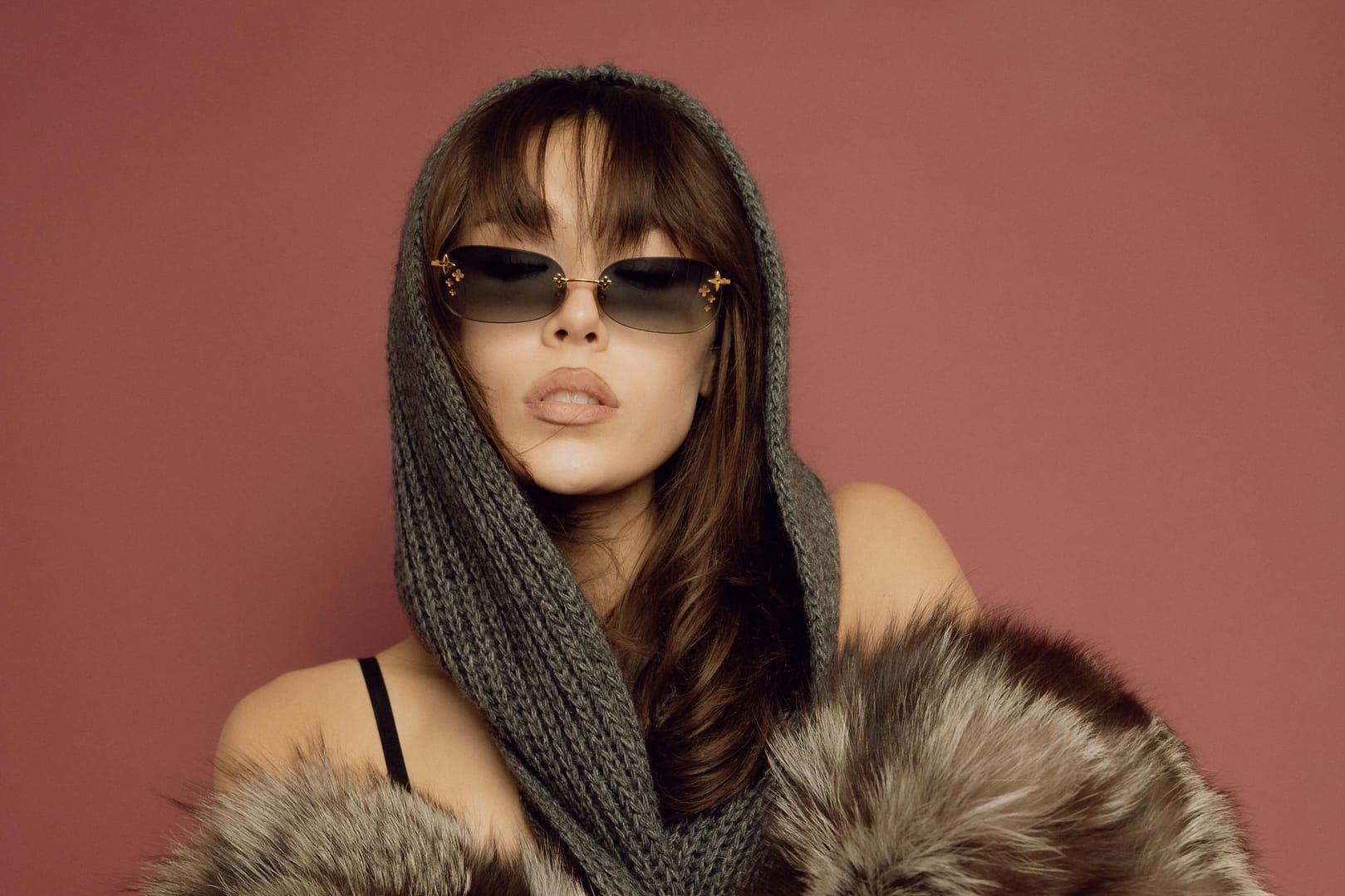 Woman wearing sunglasses and a knitted hood poses against a reddish background, draped in a faux fur garment
