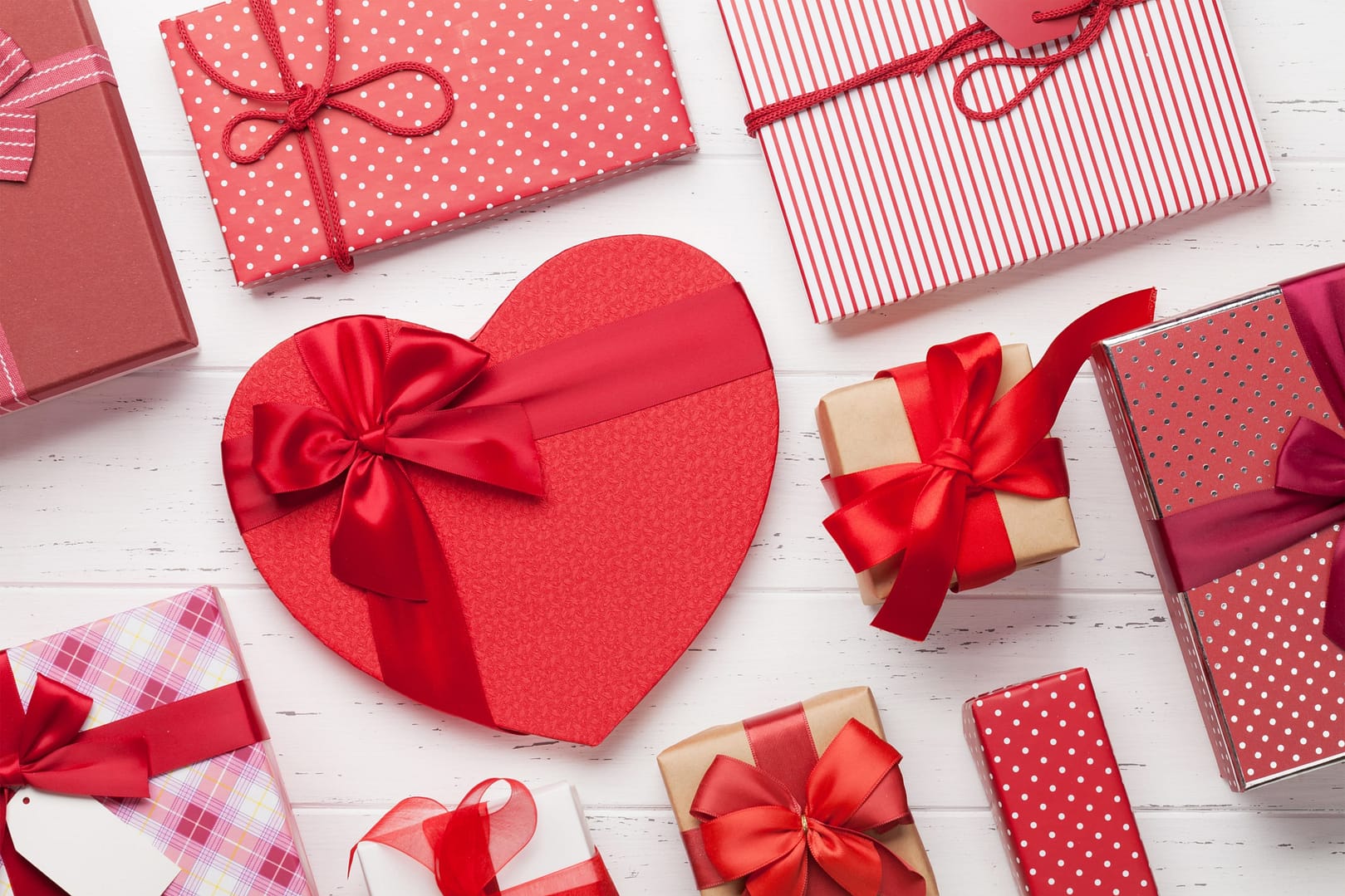 A variety of wrapped gifts, including a heart-shaped box, are decorated with red ribbons and patterned paper, arranged on a white wooden surface