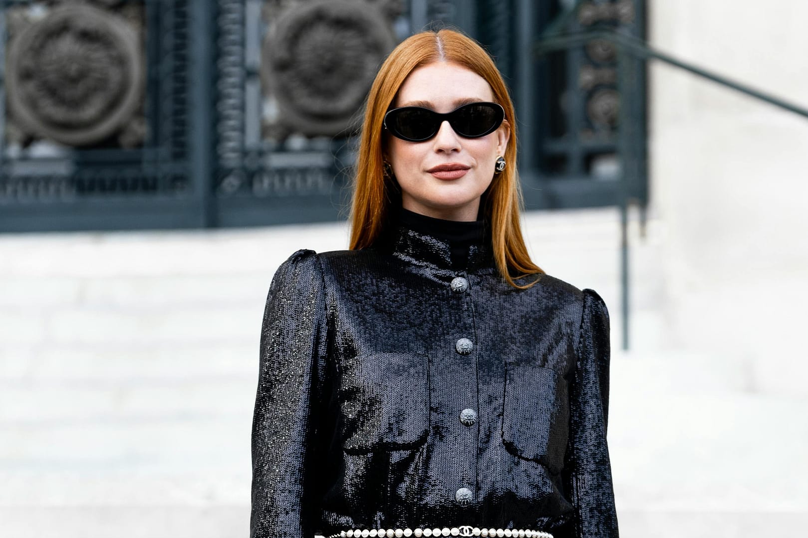 Woman looking very chic in a black sequined outfit and stylish sunglasses, holds a small black handbag while posing confidently on steps before ornate metal doors