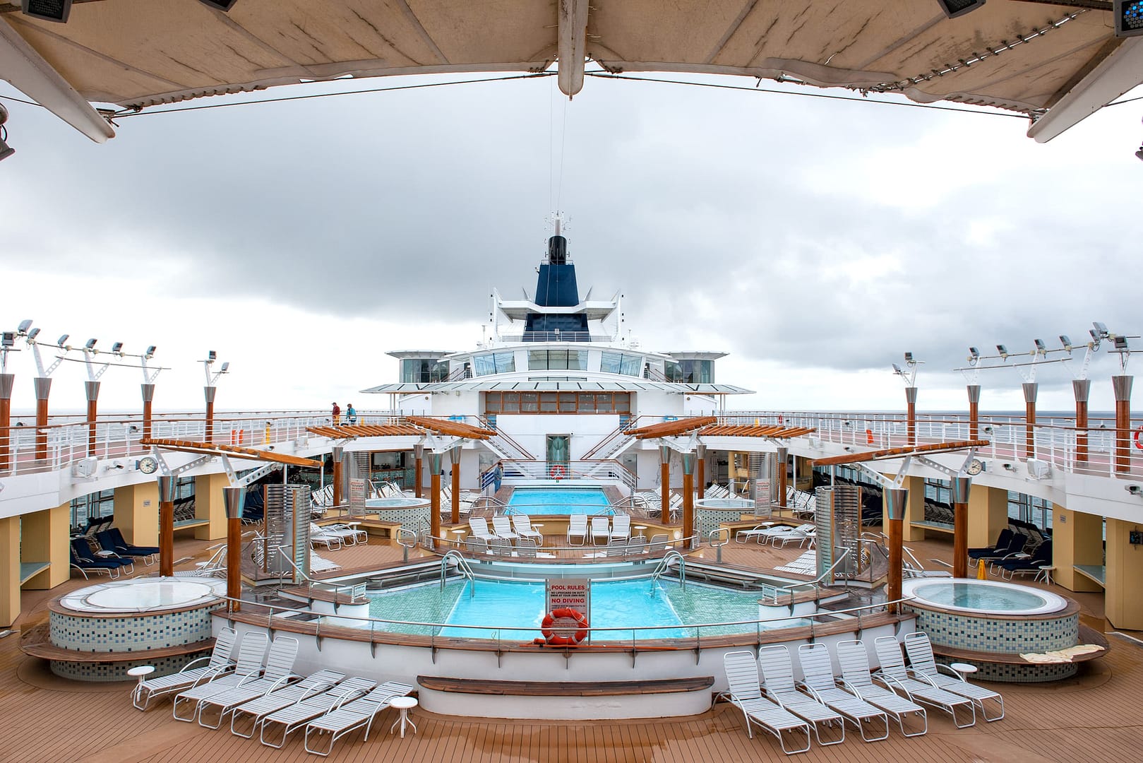 Cruise ship deck with multiple pools, hot tubs, and loungers. Overcast sky in the background