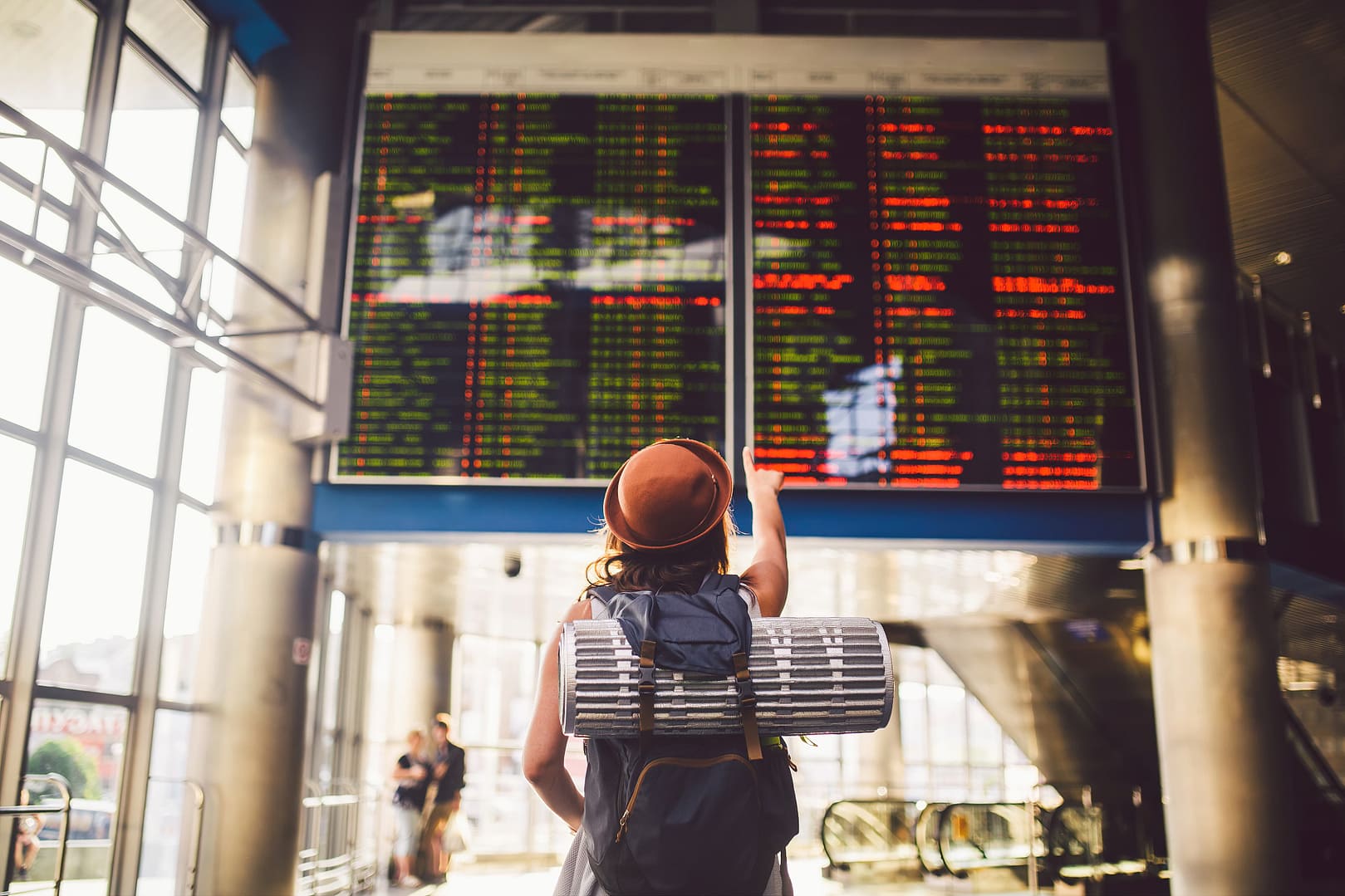 Woman in an airport station looking at a departure board while holding a suitcase and wearing a backpack and a hat