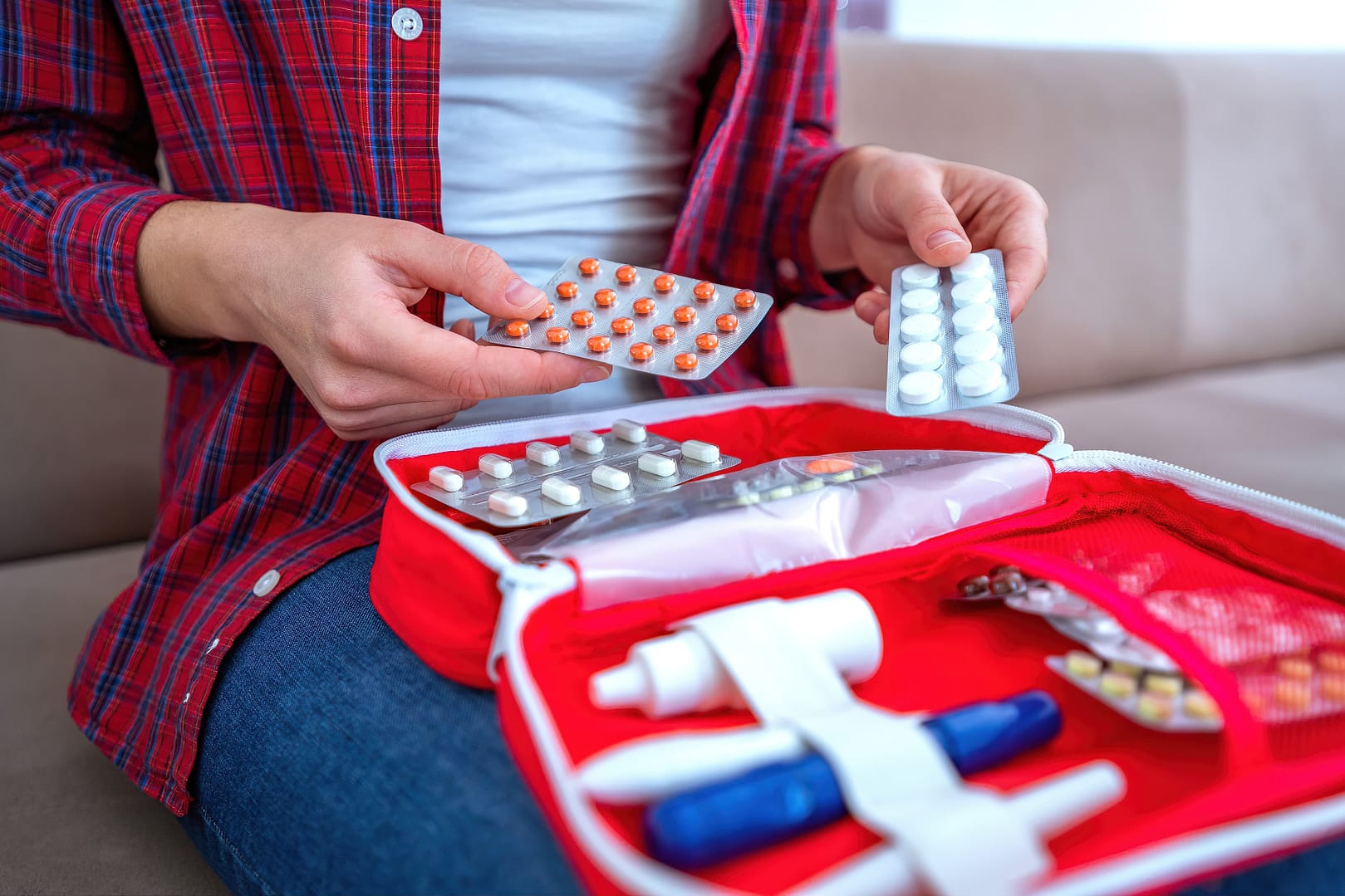 A person holding medication blister packs over an open first aid kit filled with various medical supplies