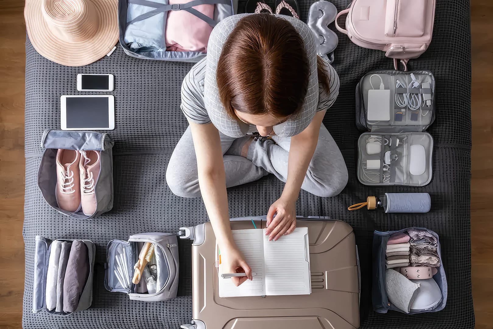 A woman is sitting on a bed with her luggage making a packing list for her trip