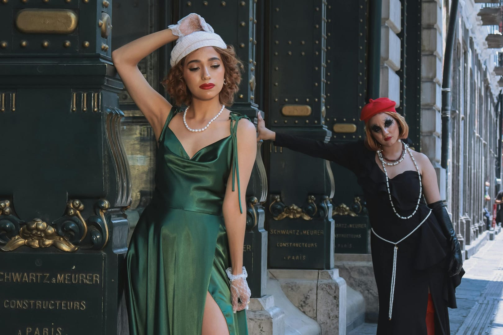 Two women pose stylishly on a city street. One wears a green dress, white gloves and white beret while the other complements her black and red outfit with a chic red beret. They stand in front of ornate doors