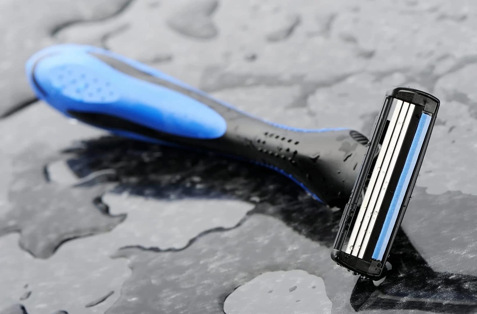 A silver razor blade resting on a damp surface, with droplets of water visible in the background