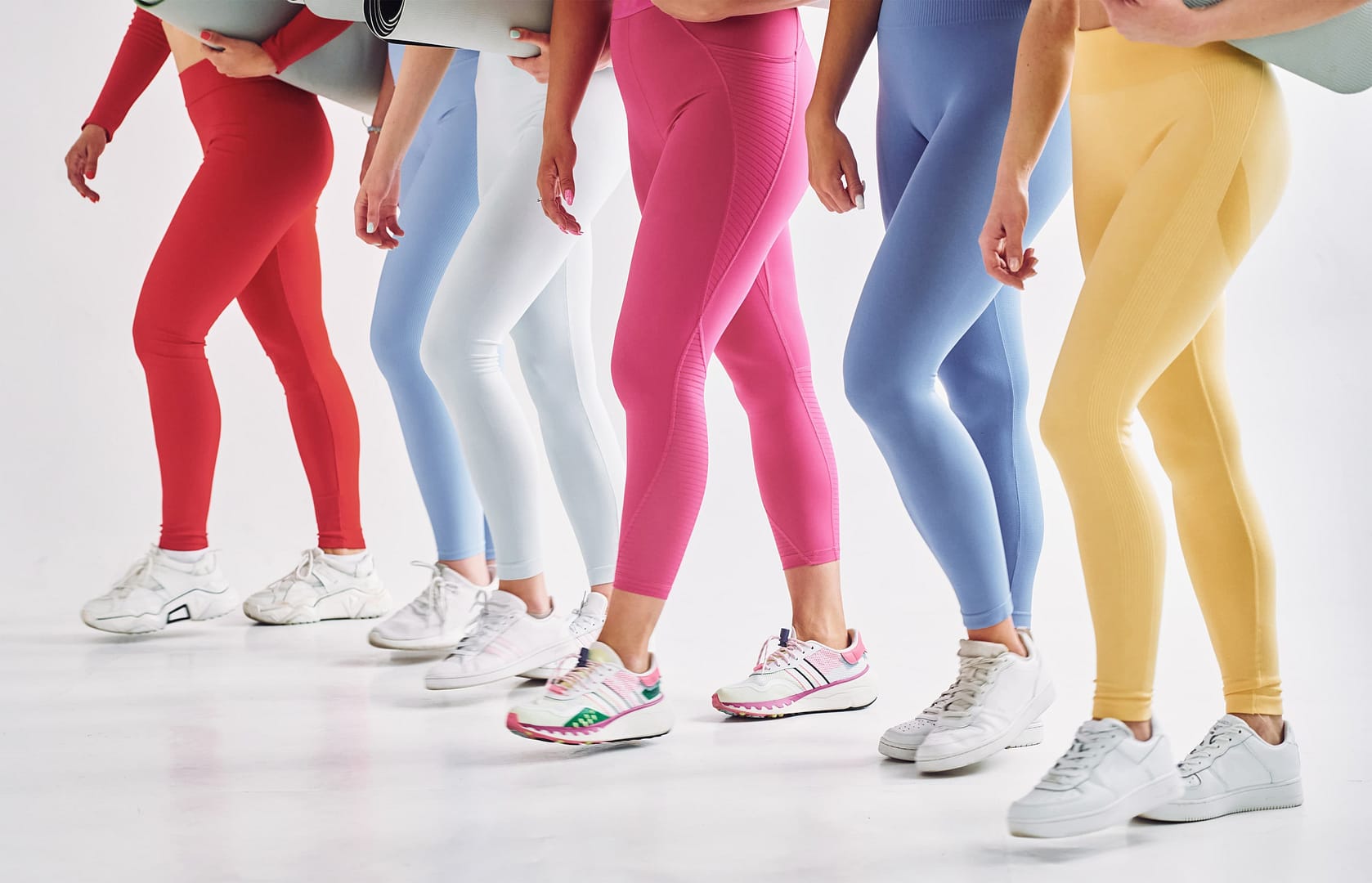 A group of people in vibrant leggings and white sneakers, clutching rolled yoga mats, stands confidently against a pristine white background