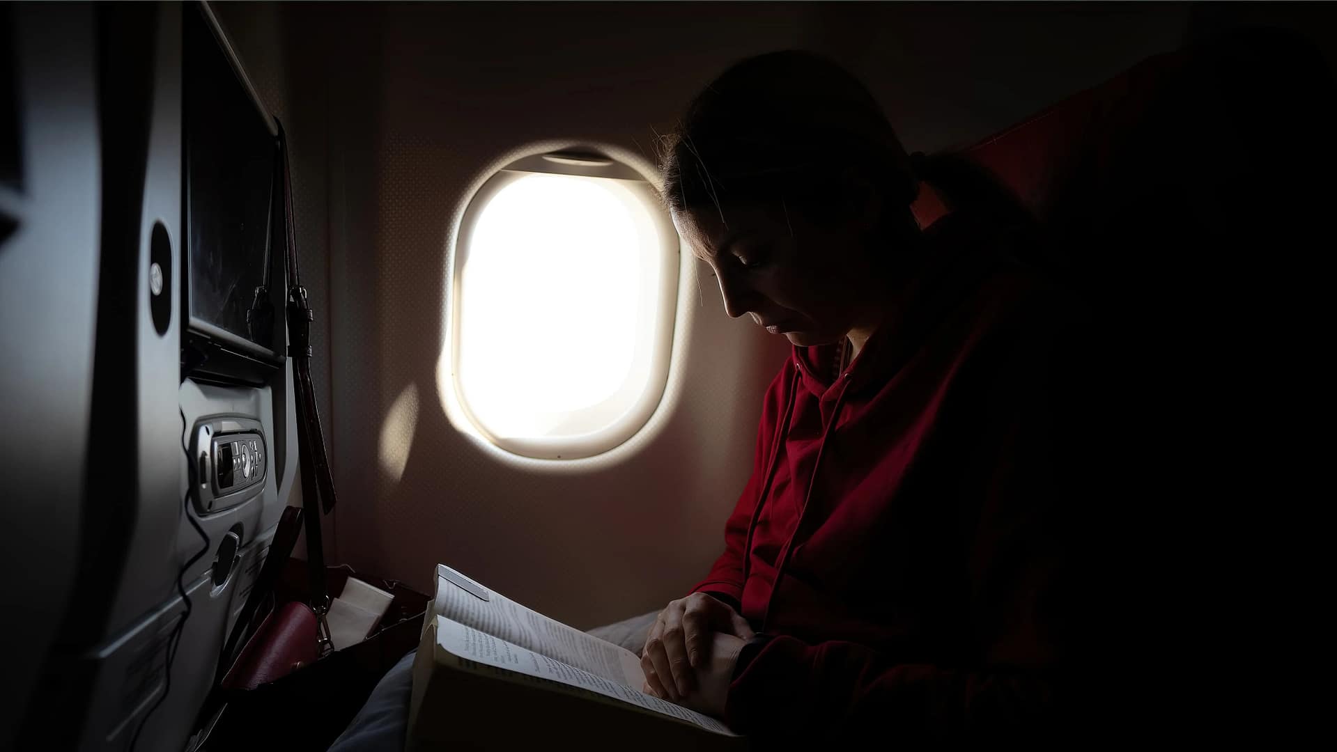 A blonde woman in a red hoodie reads a book by the light of an airplane window