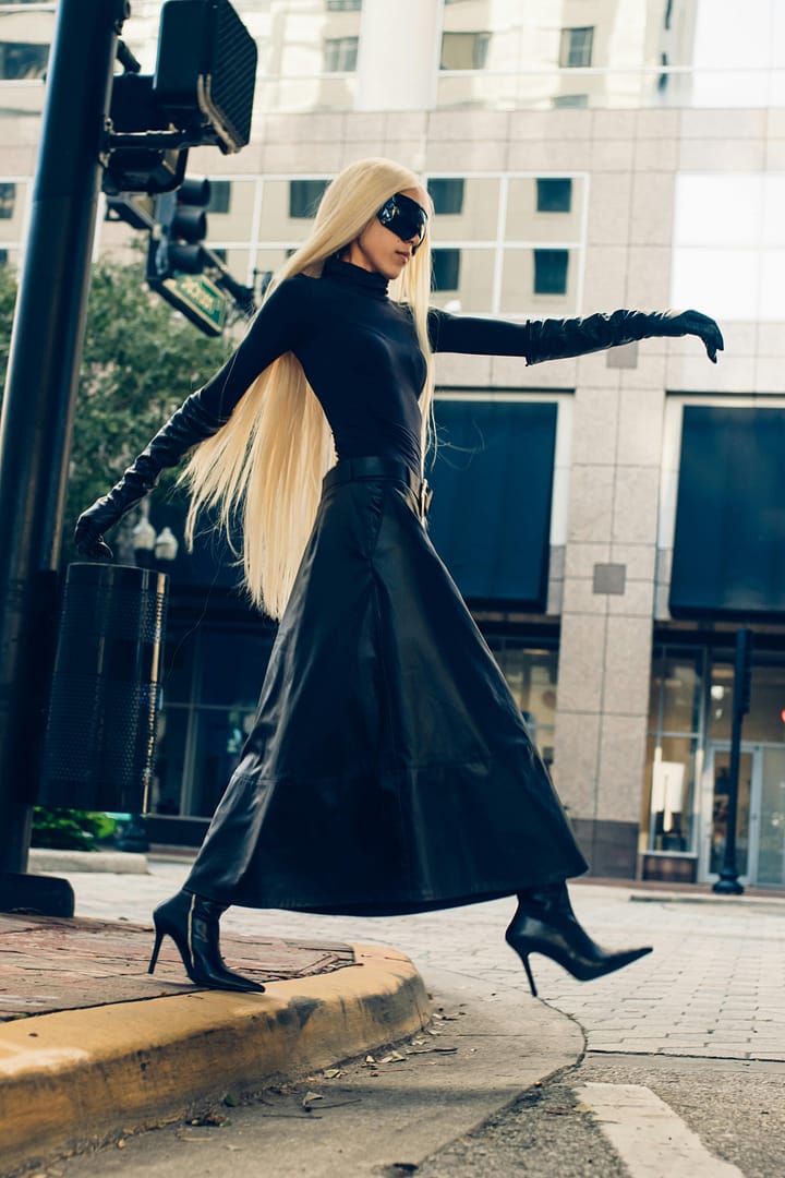 Woman in a stylish black outfit and long blonde hair strides confidently across an urban street, their chic sunglasses adding an air of mystery while high heels click on the pavement