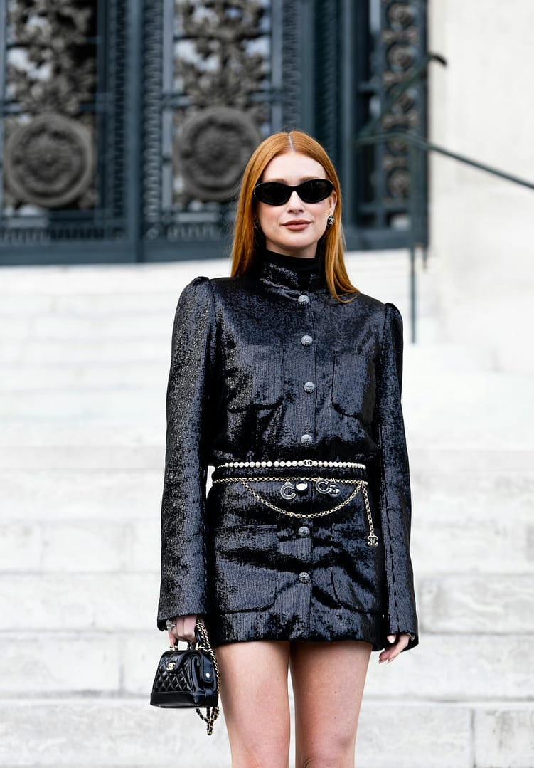 Woman looking very chic in a black sequined outfit and stylish sunglasses, holds a small black handbag while posing confidently on steps before ornate metal doors