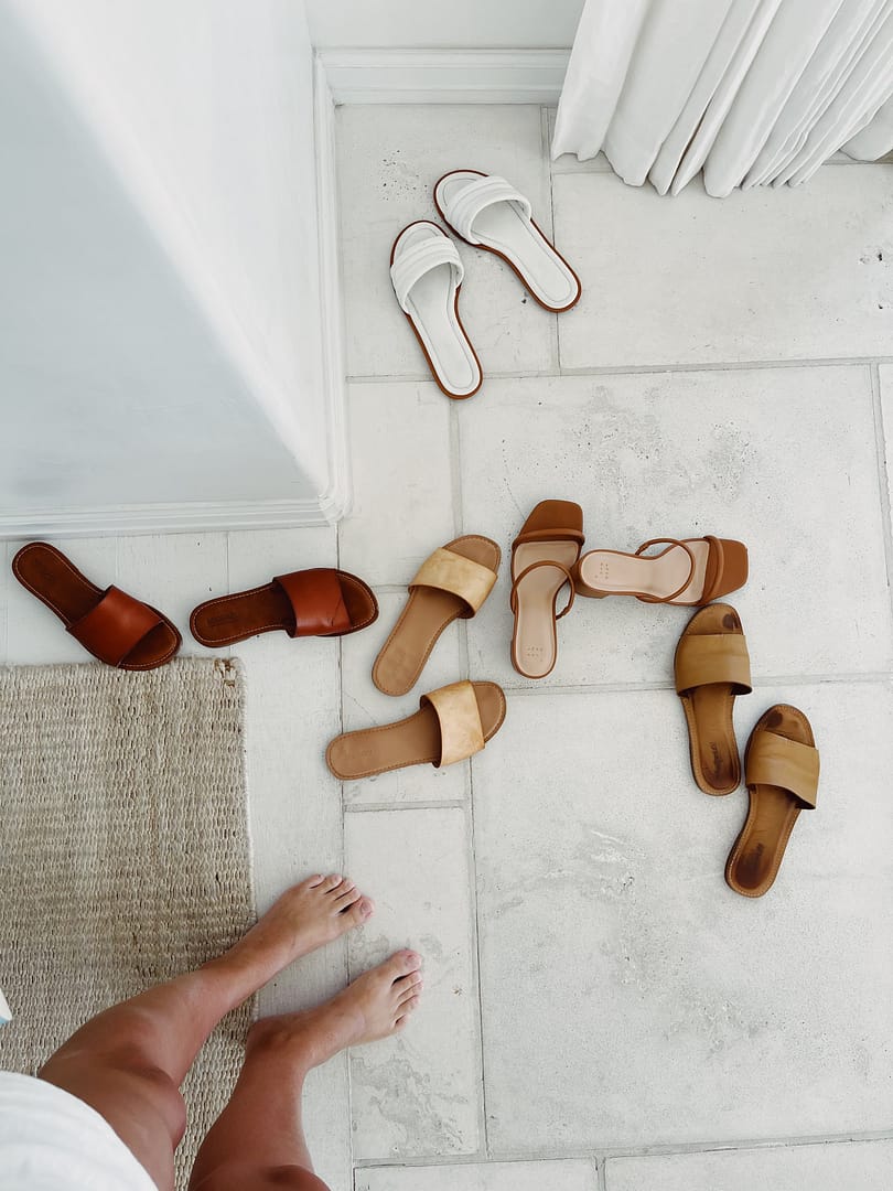 Woman stands barefoot on a tiled floor, encircled by an array of stylish brown and white sandals