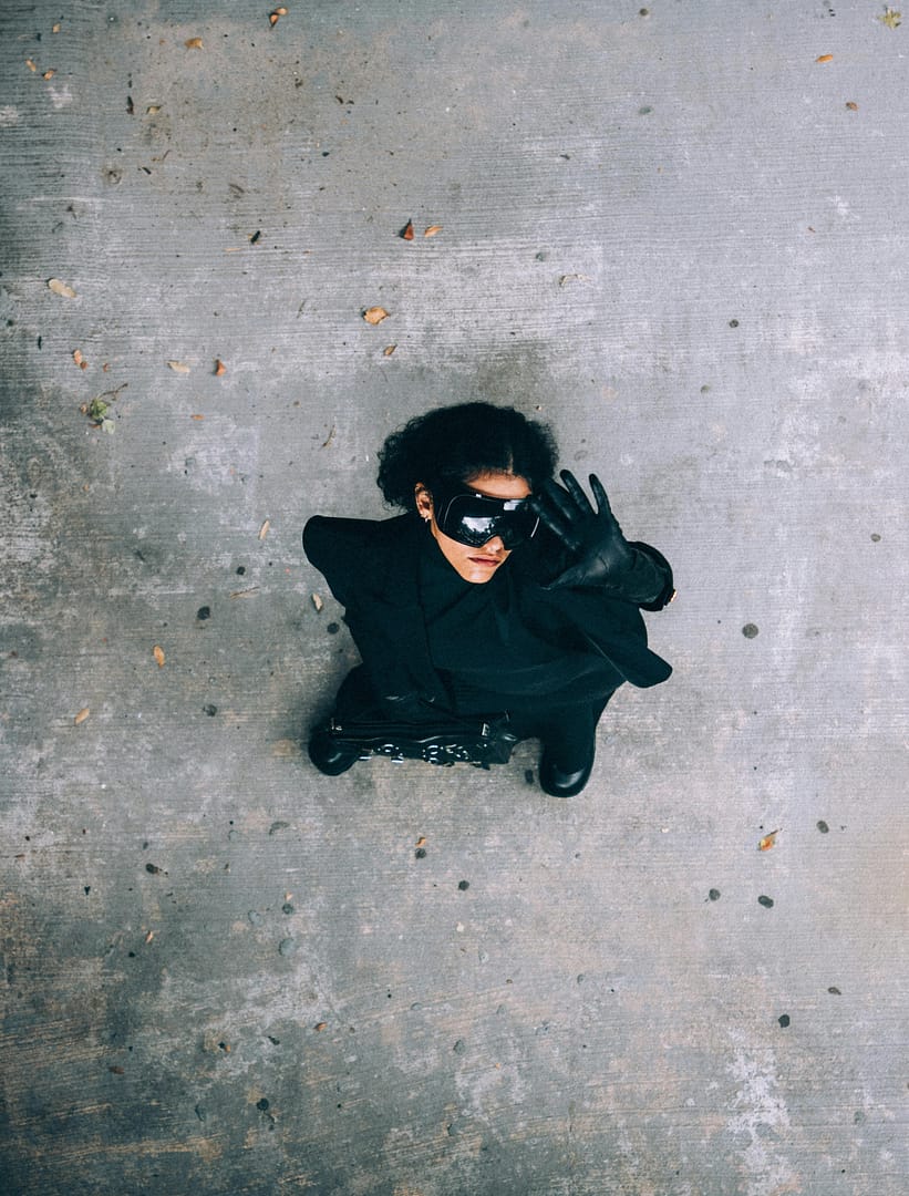 Woman dressed in black and wearing sunglasses stands on a concrete surface, looking up with one hand raised. Fallen leaves are scattered around