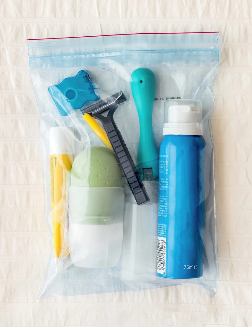 A clear plastic ziplock airline toiletry bag containing travel-sized items including a razor, toothbrush, and various bottles on a textured background