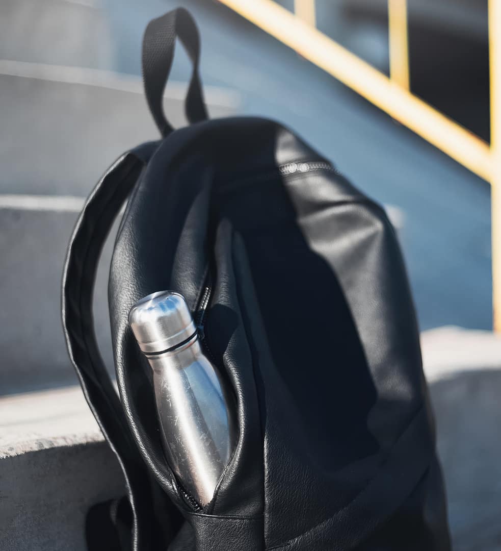 A black backpack resting on a concrete step with a stainless steel water bottle peeking out from an open side pocket